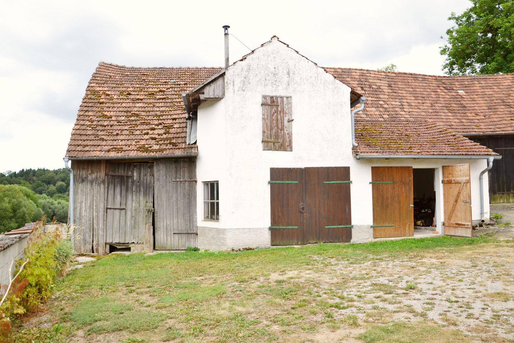 Ländliches Anwesen mit weiß getünchtem Haus, Holztüren und Ziegeldach in grüner Umgebung.
