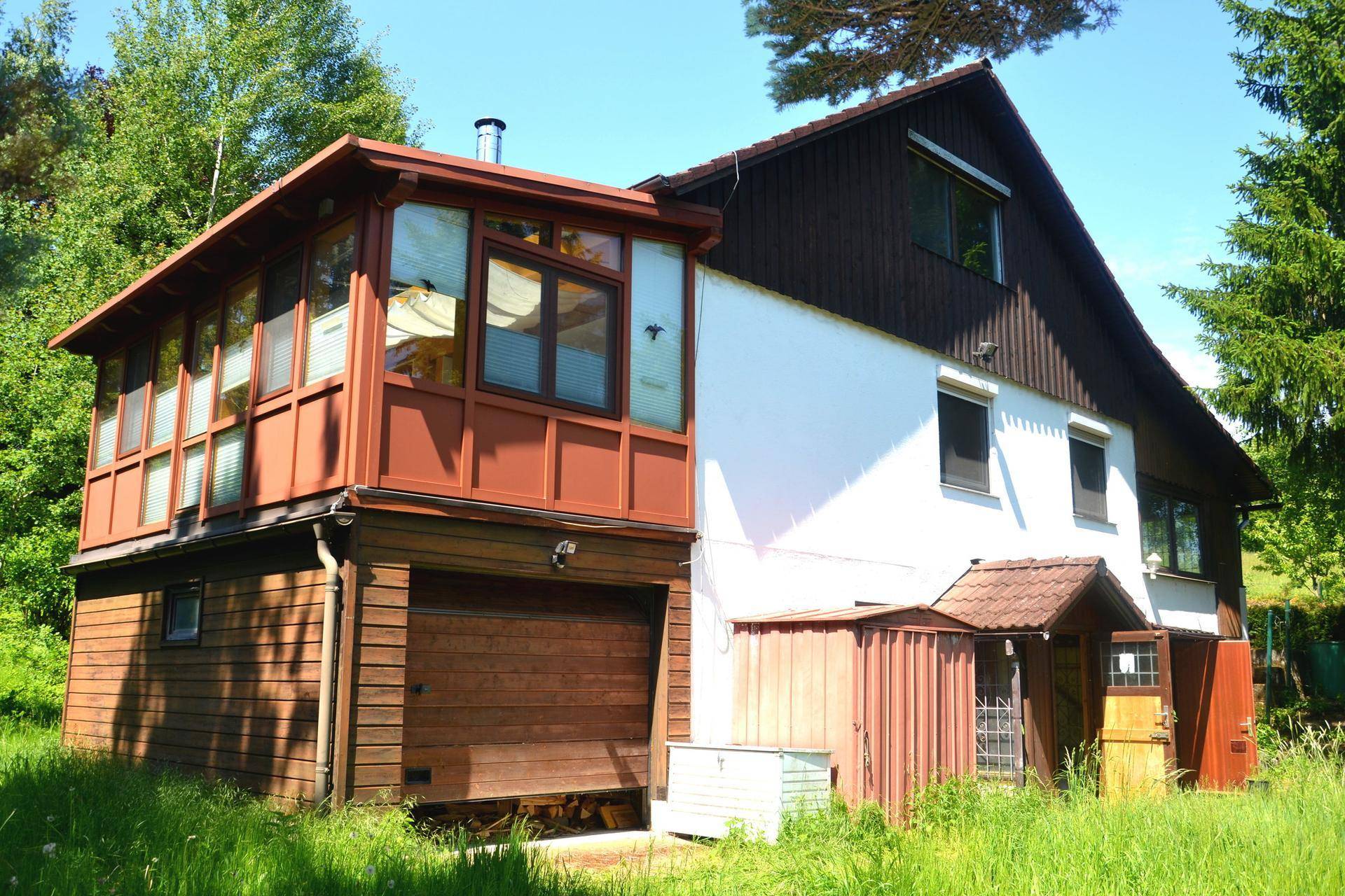 Hausfassade mit einem hölzernen Anbau, der als Wintergarten dient, und einer Garage.