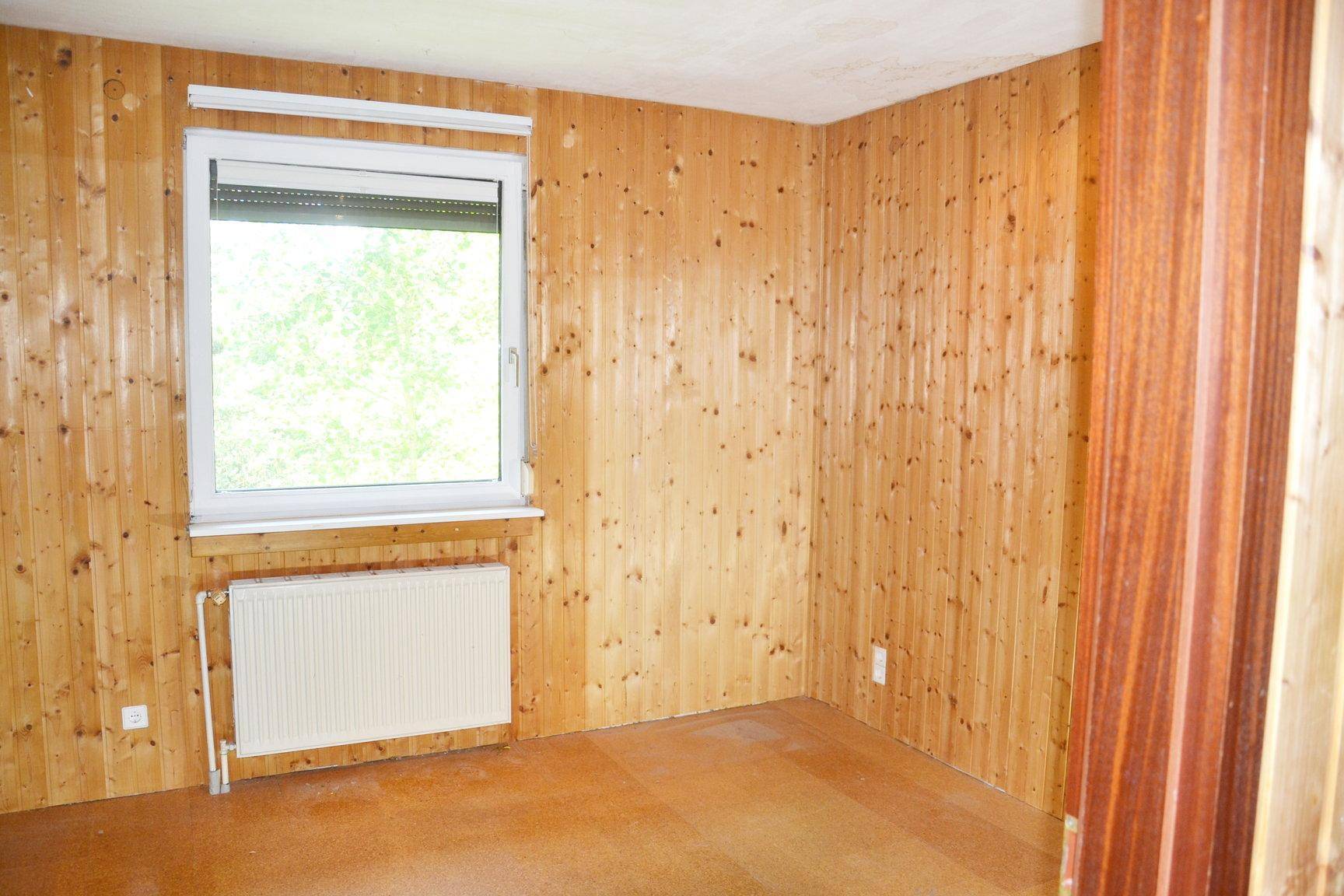 Schlafzimmer mit Holzvertäfelung, Heizkörper und Fenster mit Blick ins Grüne.