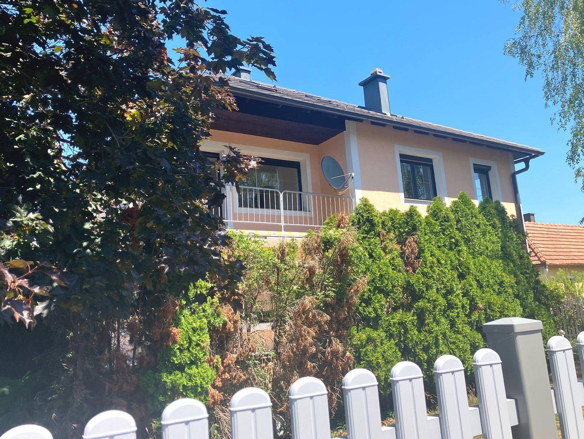 Hausfassade mit Balkon, Satellitenschüssel und grüner Bepflanzung vor einem weißen Zaun.