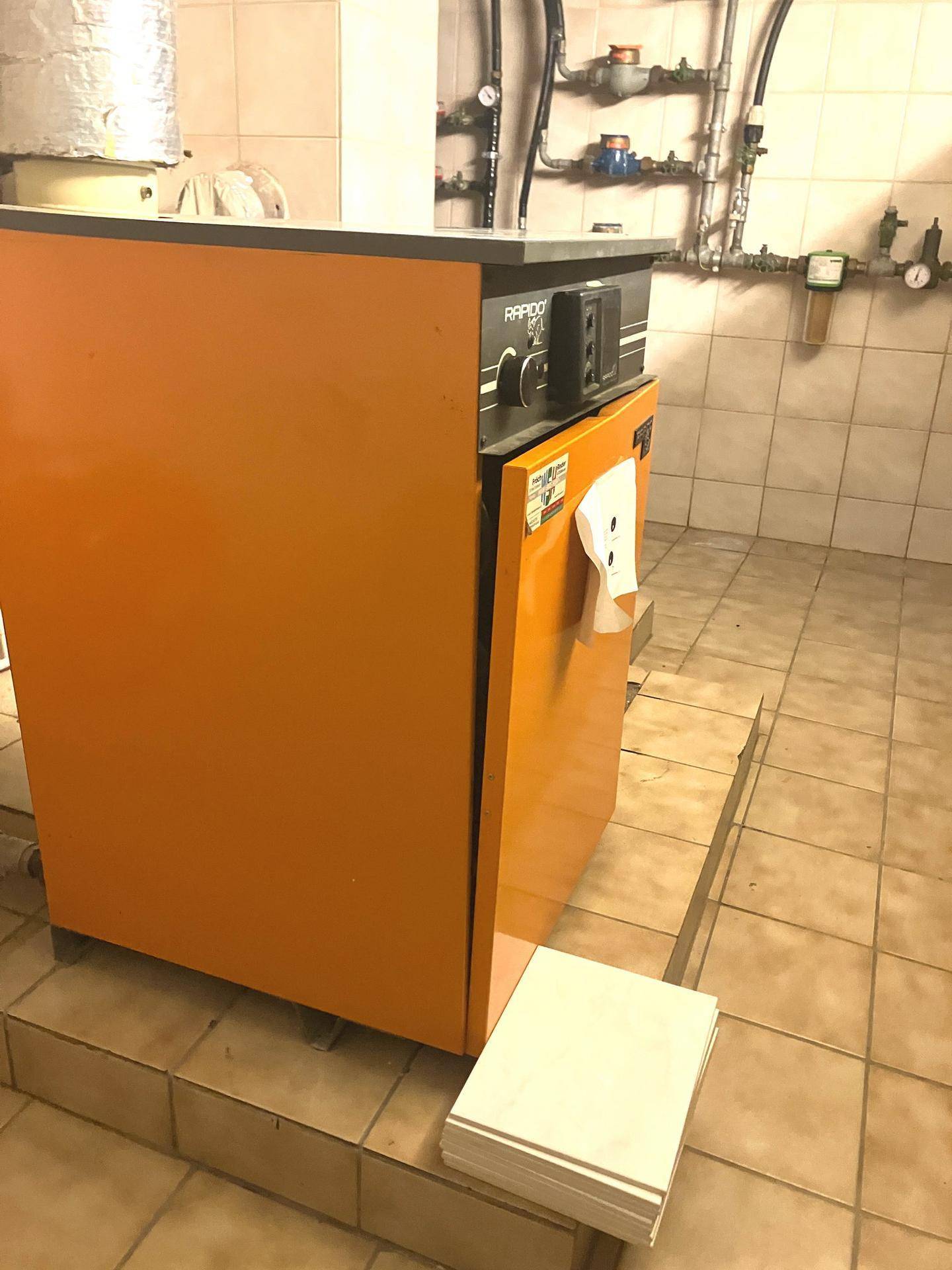 Heizungsraum mit orangefarbenem Heizkessel und gefliesten Wänden, dient der Wärmeversorgung des Hauses.