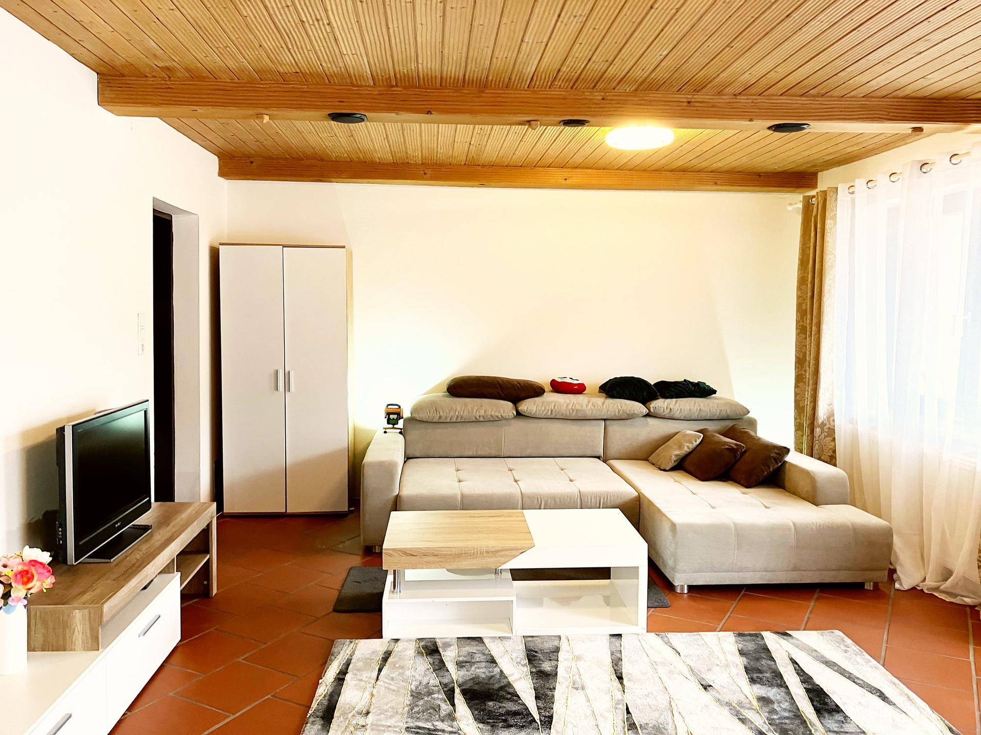 Helles Wohnzimmer mit Ecksofa, Holzbalkendecke und Terrakotta-Fliesenboden.