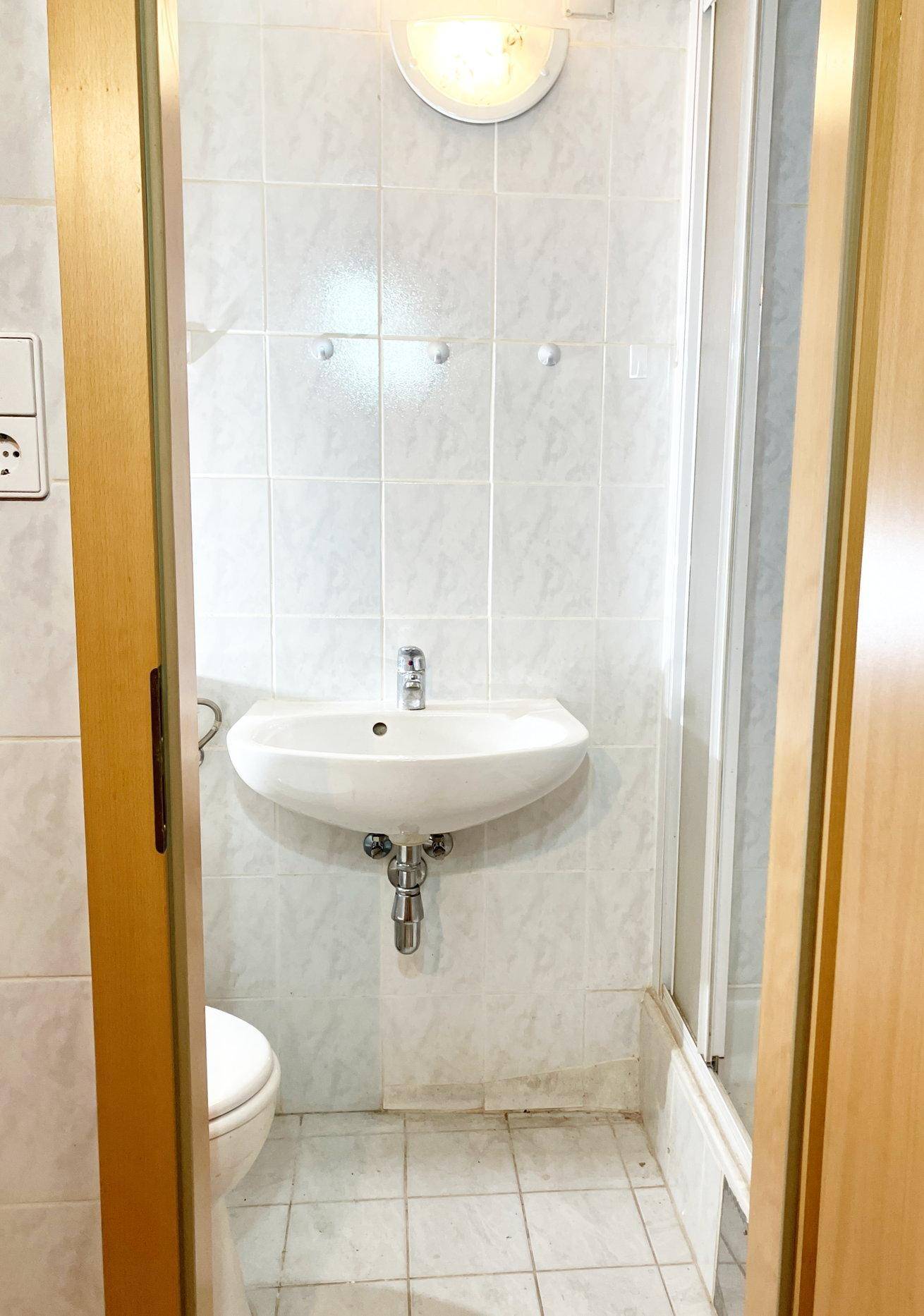 Kleines Badezimmer mit weißen Fliesen, Waschbecken und Toilette, Duschkabine teilweise sichtbar.