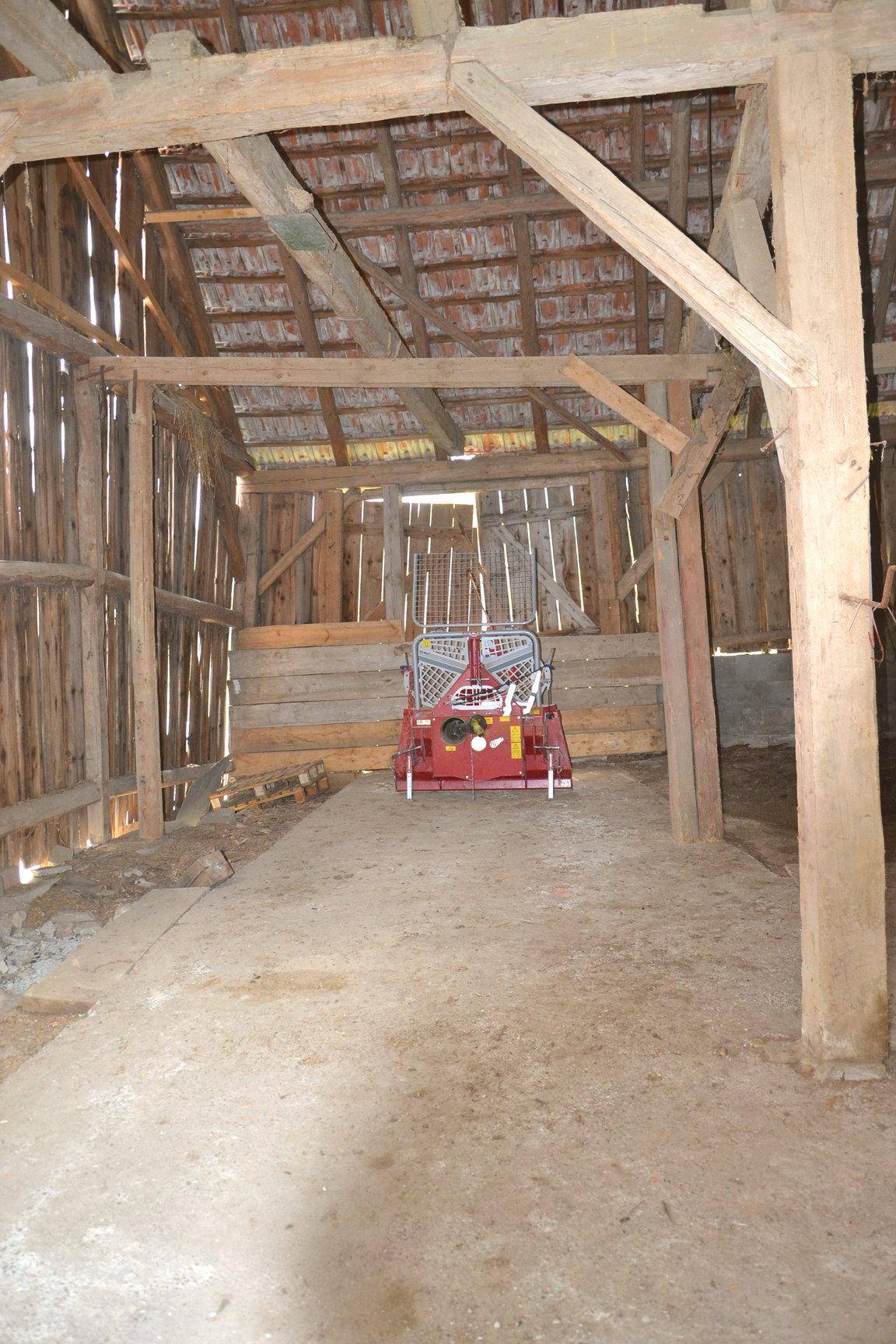 Innenansicht einer Scheune mit Holzbalken, Ziegeldach und einem landwirtschaftlichen Gerät.