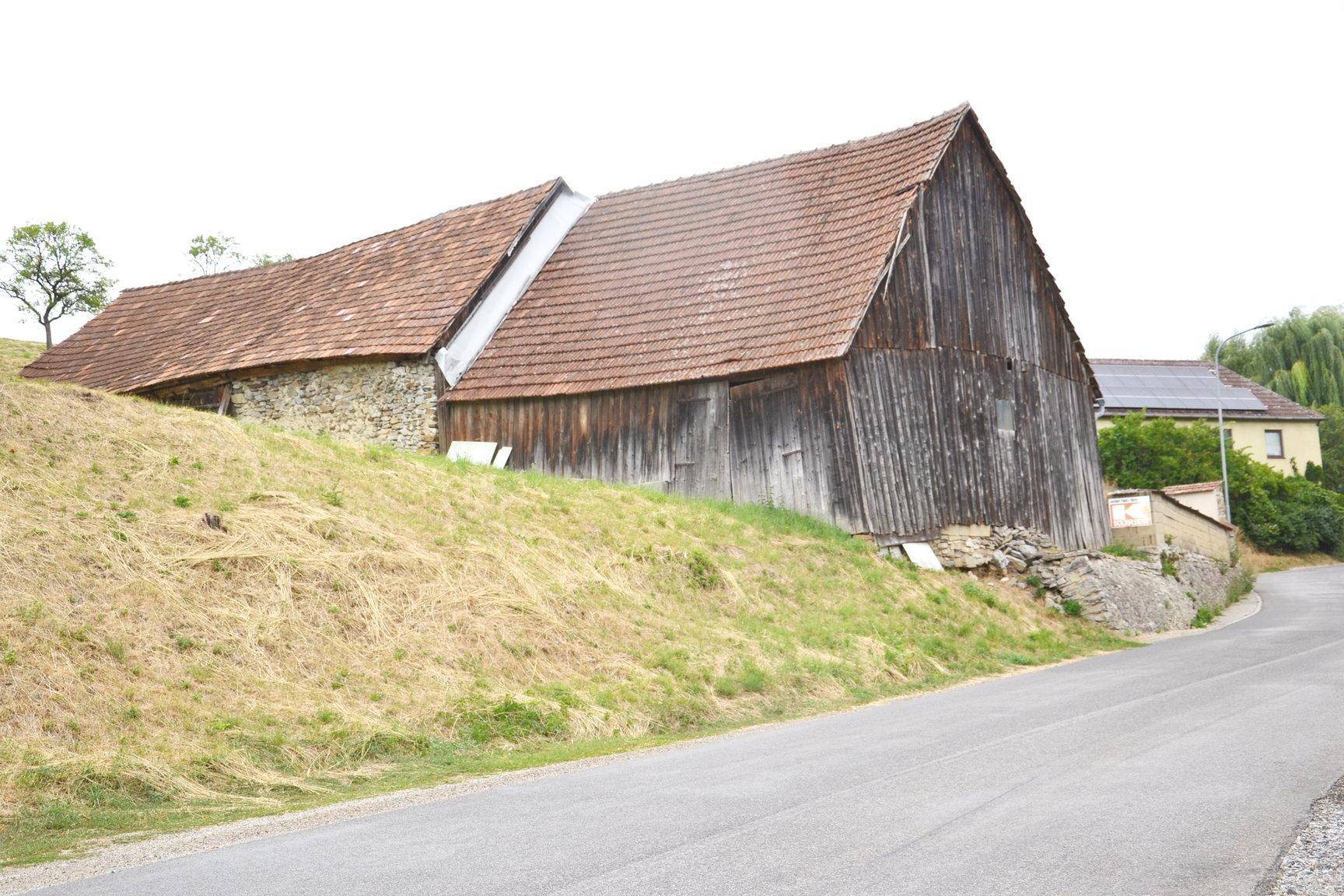 Altes Holzgebäude mit Ziegeldach an einem grasbewachsenen Hang neben einer Straße.