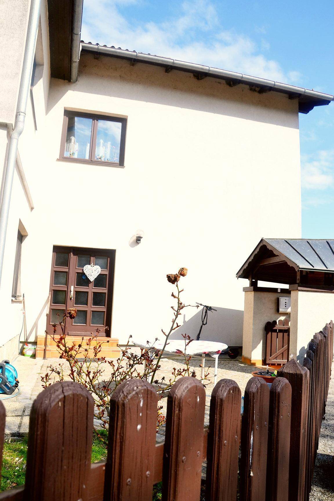 Hausfassade mit Eingangstür, Fenster und kleinem Holzzaun, umgeben von spärlicher Vegetation.