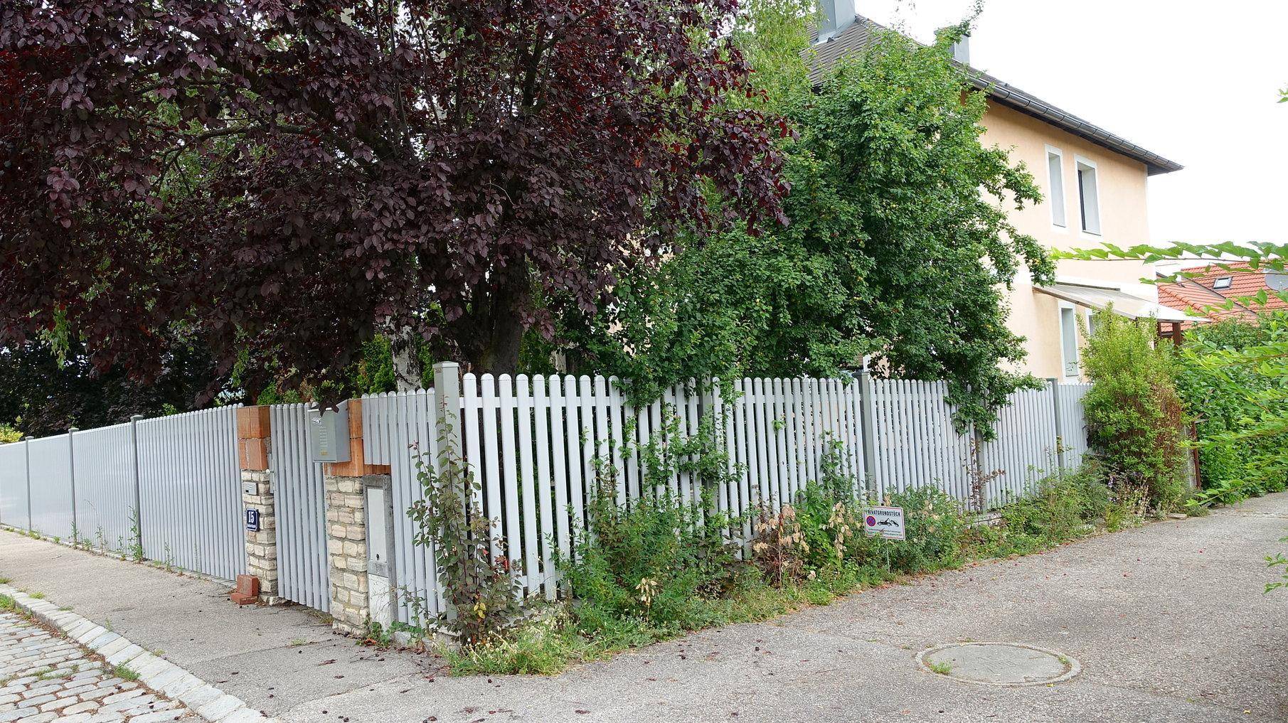 Hausansicht von der Straße mit einem weißen Zaun und üppiger Bepflanzung.