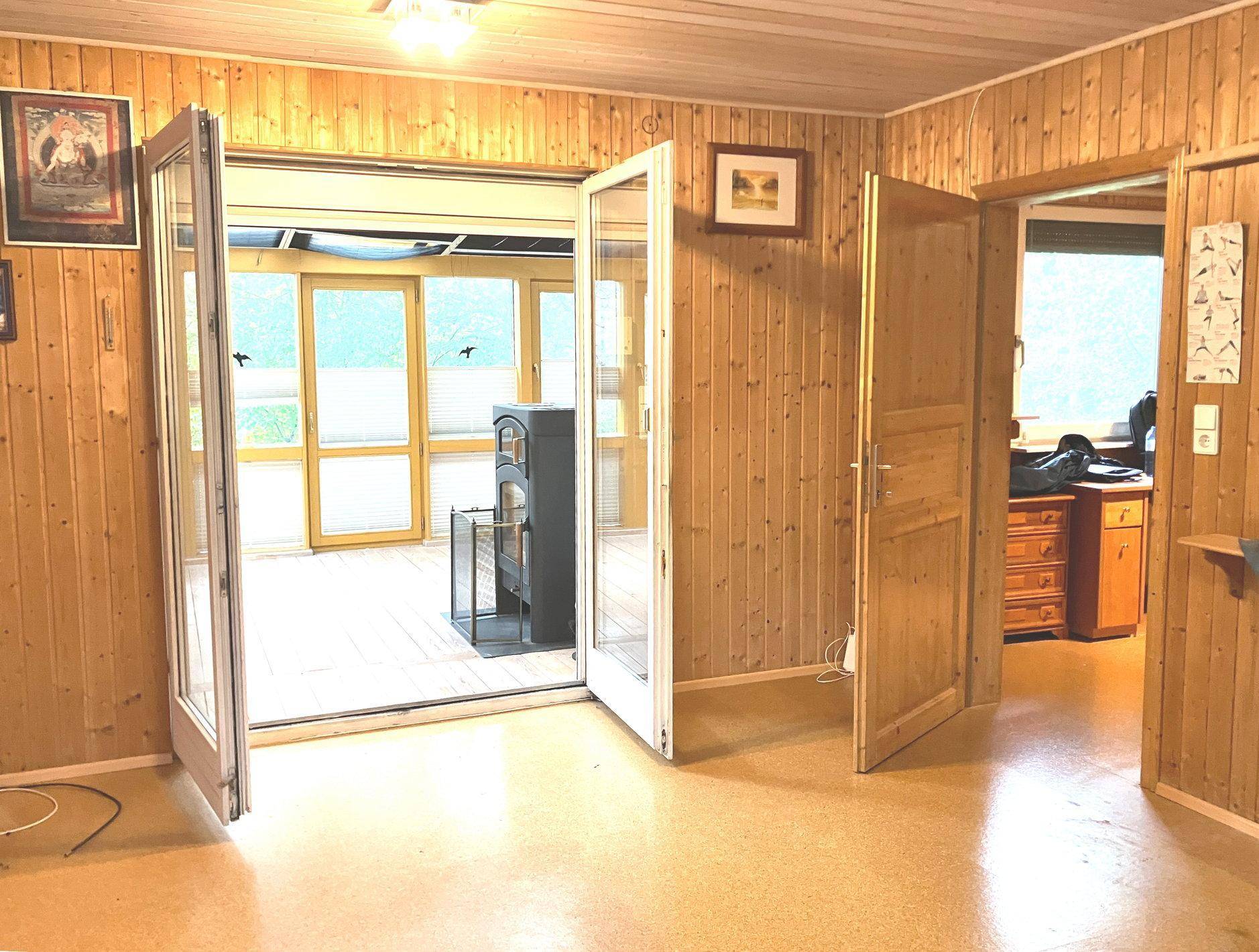 Wohnbereich mit Holzvertäfelung und Zugang zu einem hellen Wintergarten mit Kaminofen.