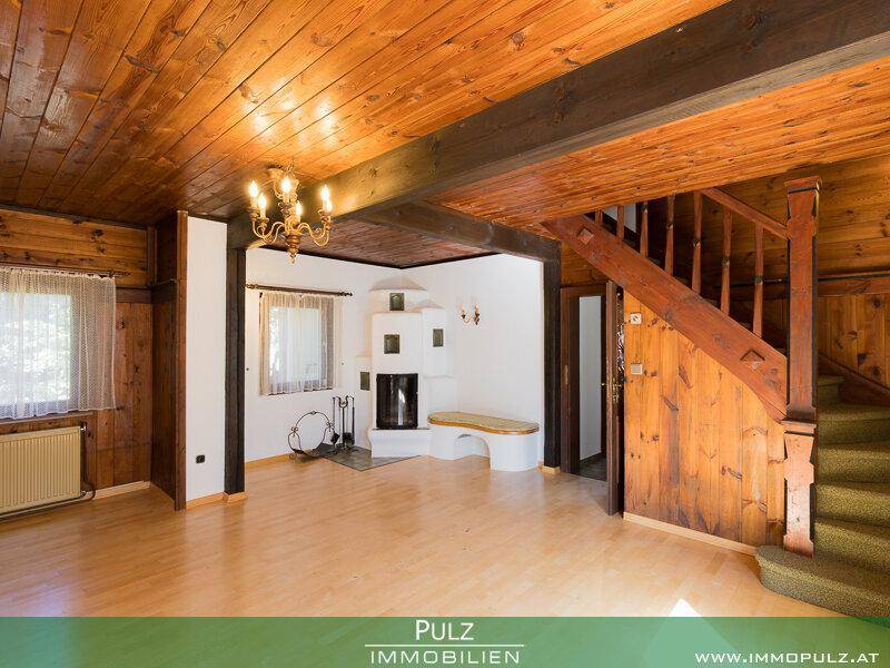 Gemütliches Wohnzimmer mit Holzbalkendecke, Holzwänden, Kachelofen und einer Holztreppe.