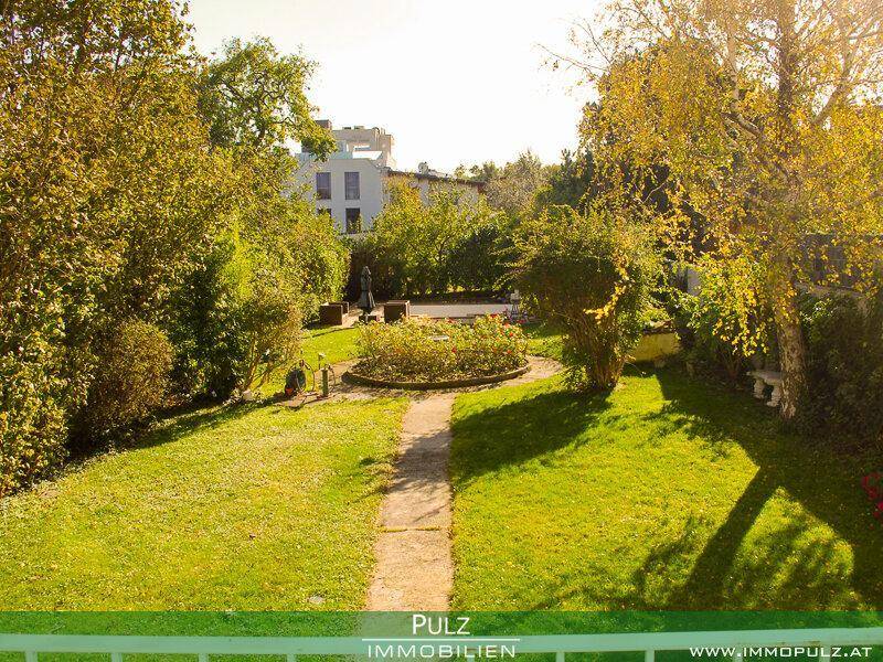 Weitläufiger Garten mit Rasen, Bäumen und einem Rundbeet, im Hintergrund ein Gebäude.