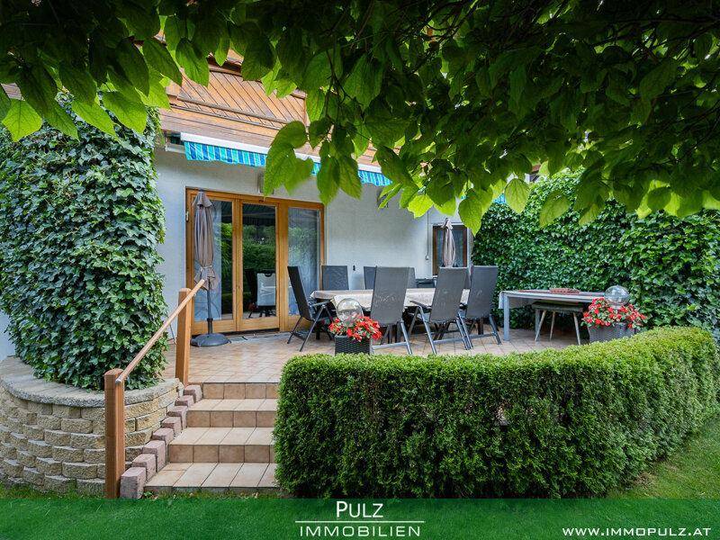 Große Terrasse mit Esstisch und Stühlen, umgeben von hohen Hecken und grüner Bepflanzung.