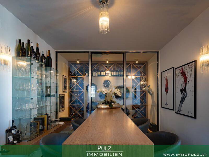 Exklusiver Weinkeller mit Verkostungsbereich, Holztheke und beleuchteten Weinregalen hinter Glas.