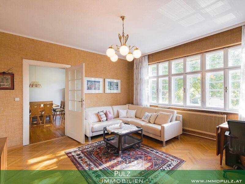 Helles Wohnzimmer mit beiger Couch, Parkettboden und Zugang zu einem angrenzenden Essbereich.