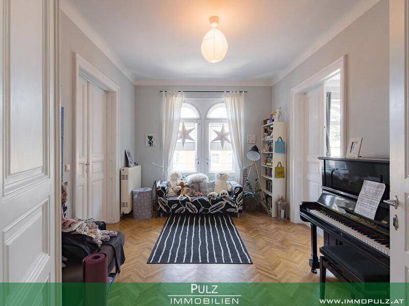 Helles Zimmer mit Parkettboden, großem Fenster und einem Klavier, ideal als Kinder- oder Gästezimmer.