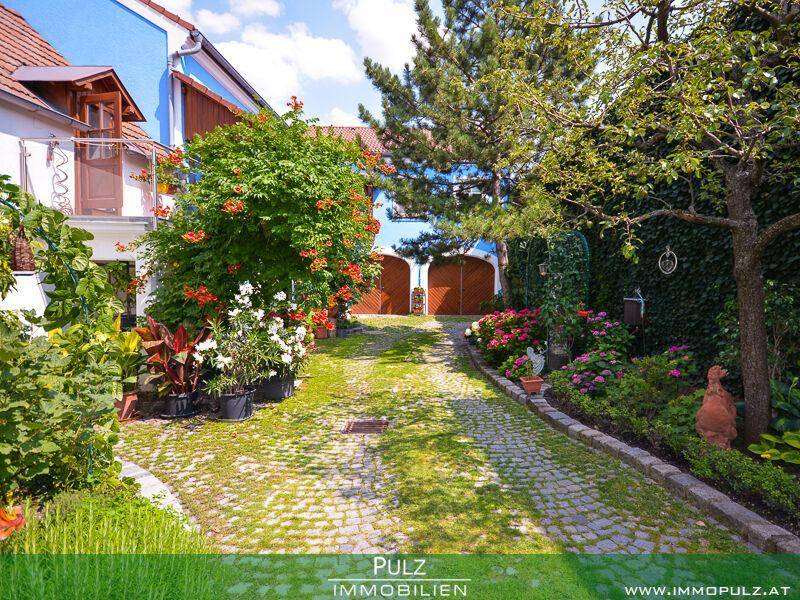 Gepflasterter Innenhof mit üppigen Blumenbeeten, Bäumen und bunten Hausfassaden unter blauem Himmel.