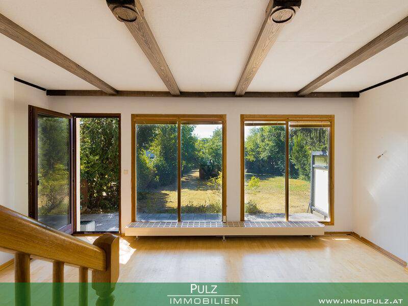 Heller Raum mit Holzboden, sichtbaren Deckenbalken und großen Fenstern mit Zugang zum Garten.