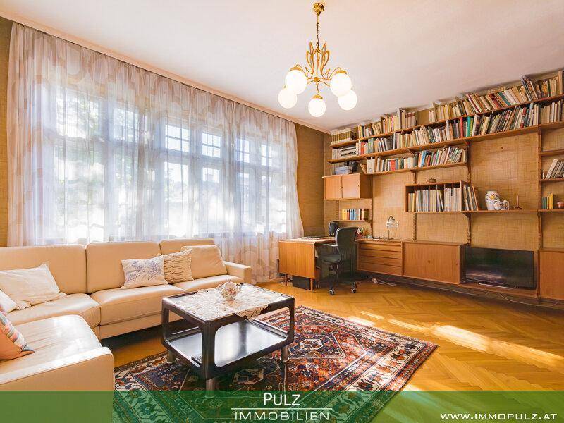 Geräumiges Wohnzimmer mit Parkettboden, großer Couch und einer Bücherwand, hell durch große Fenster.