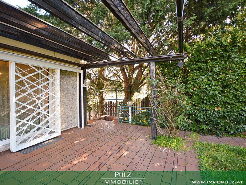 Überdachte Terrasse mit Pergola und Zugang zum Haus, umgeben von Grün.