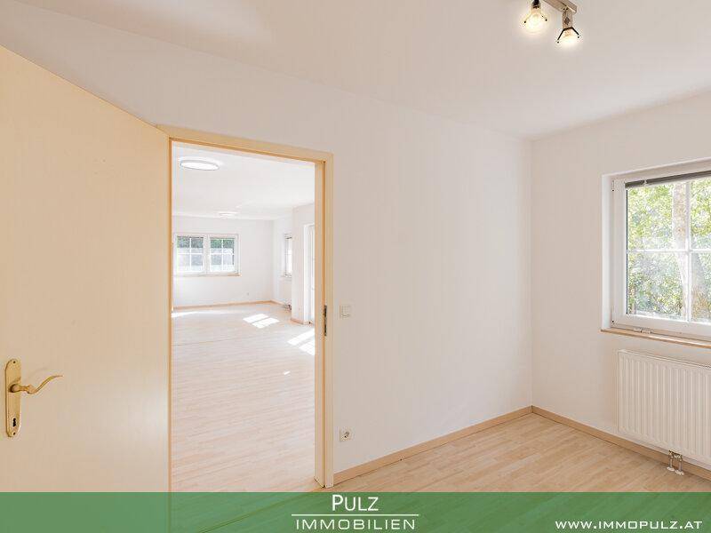 Leeres Zimmer mit Holzboden und Fenster, bietet Blick in einen weiteren hellen Raum.