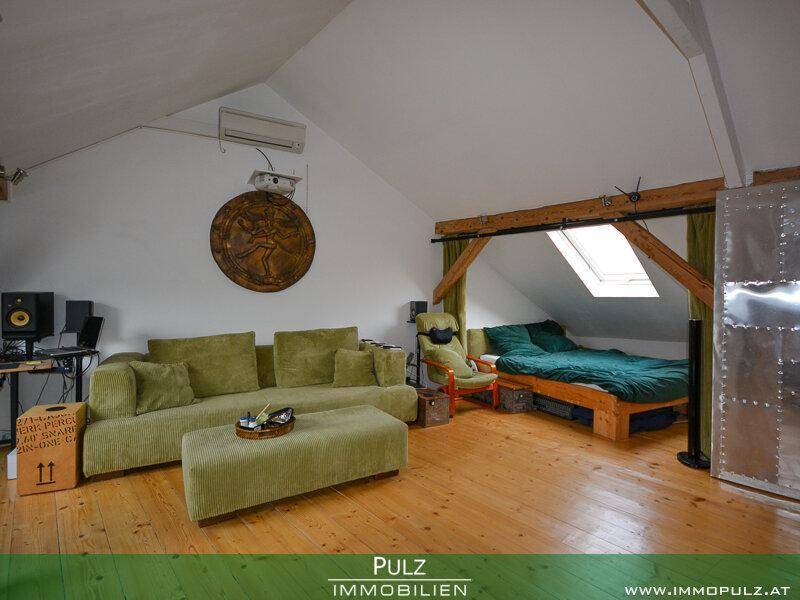 Offener Wohnbereich im Dachgeschoss mit Holzboden, Sofa und einem Bett unter dem Dachfenster.