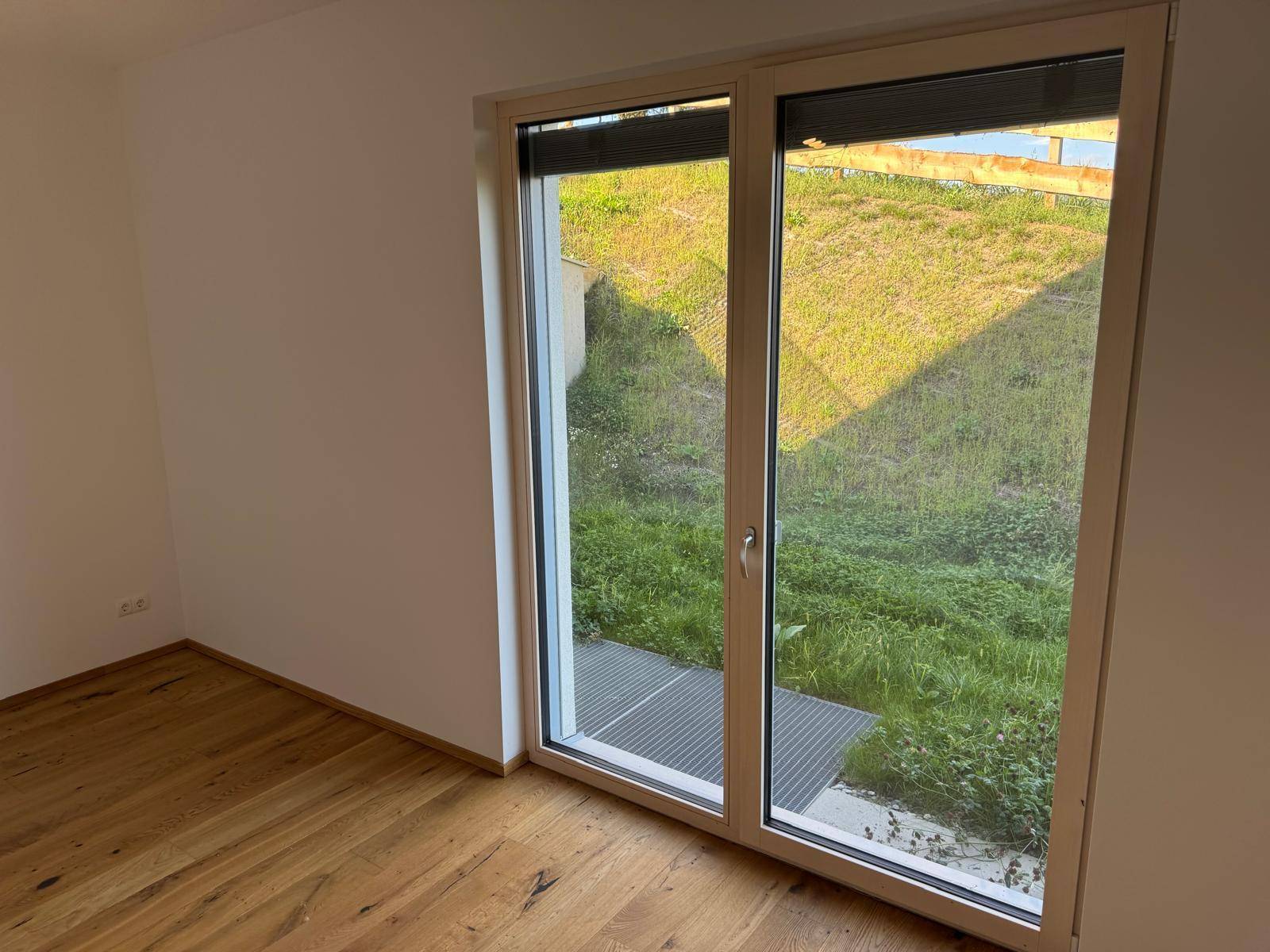 Heller Raum mit Holzboden und großer Terrassentür, die zu einem gepflasterten Außenbereich führt.