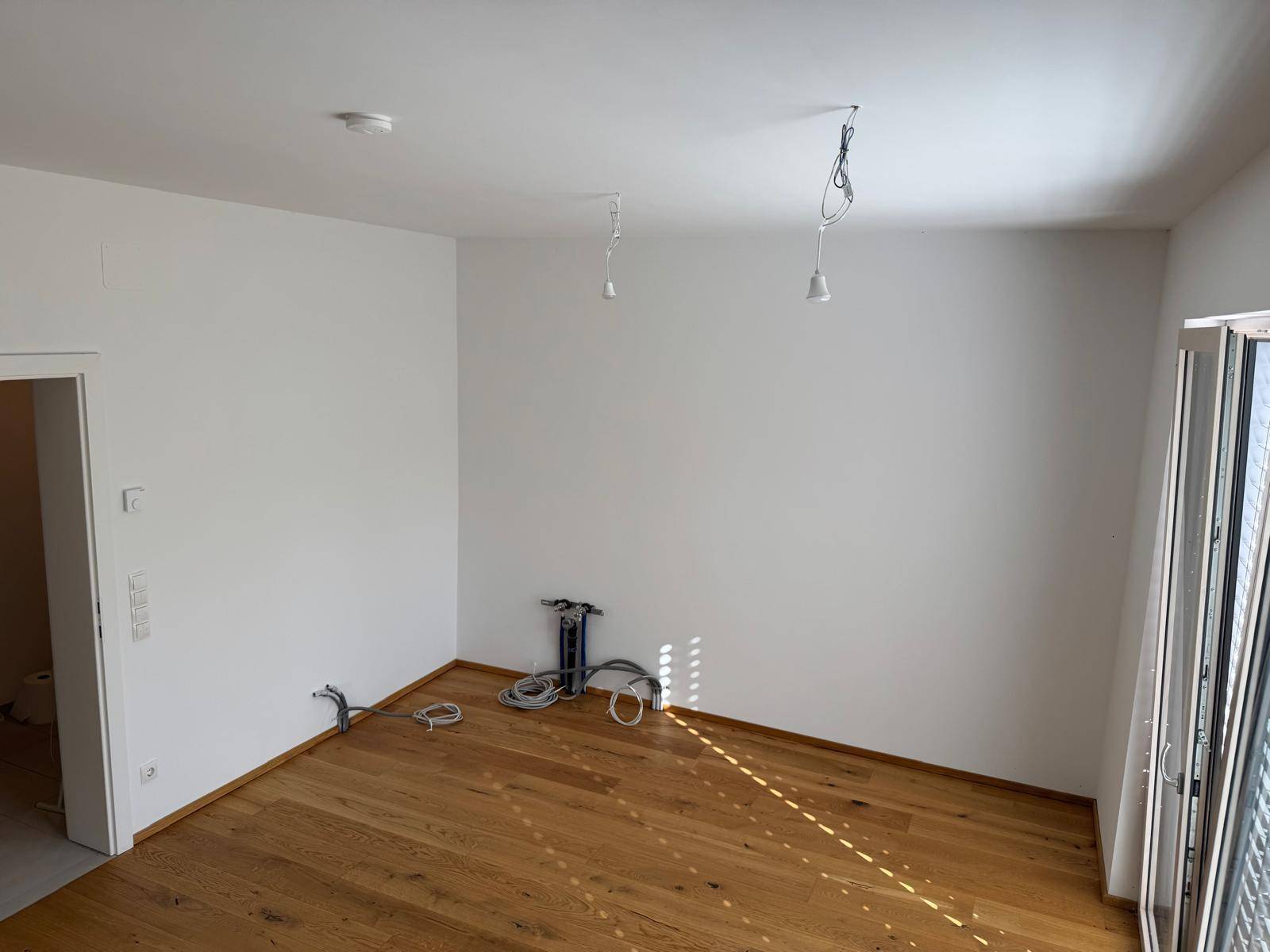 Leerer Raum mit Holzboden und vorbereiteten Anschlüssen, ideal für Wohn- oder Küchenbereich.