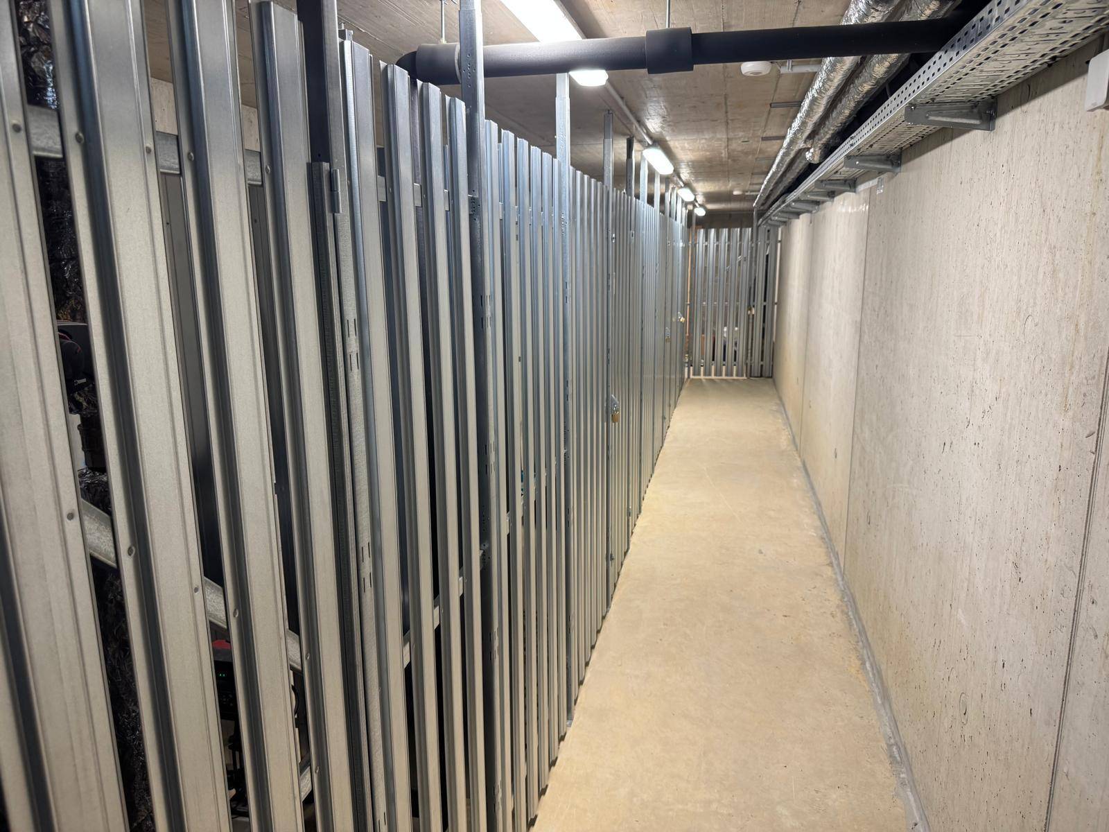 Langer Kellergang mit Metallabtrennungen für individuelle Abstellräume und Betonwänden.