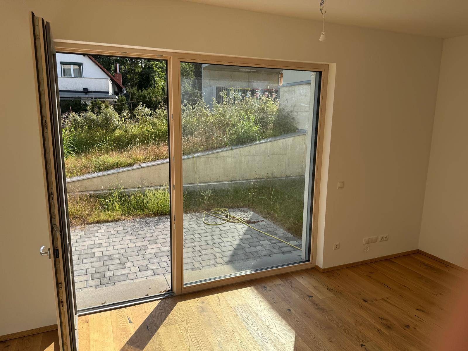 Heller Raum mit Holzboden und großer Terrassentür, die zu einem gepflasterten Außenbereich führt.