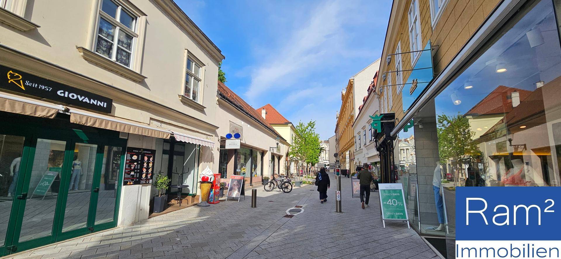 Belebtes Stadtbild mit Geschäften und Fußgängern entlang einer historischen Straße unter blauem Himmel.