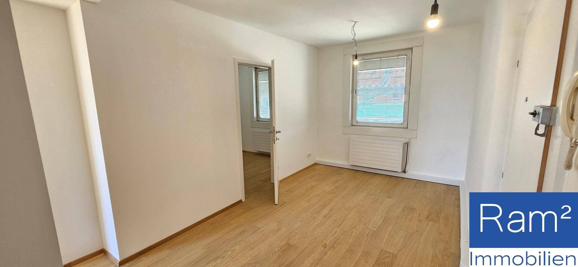 Leeres Wohnzimmer mit hellem Laminatboden und einem Fenster, das viel Tageslicht hereinlässt.