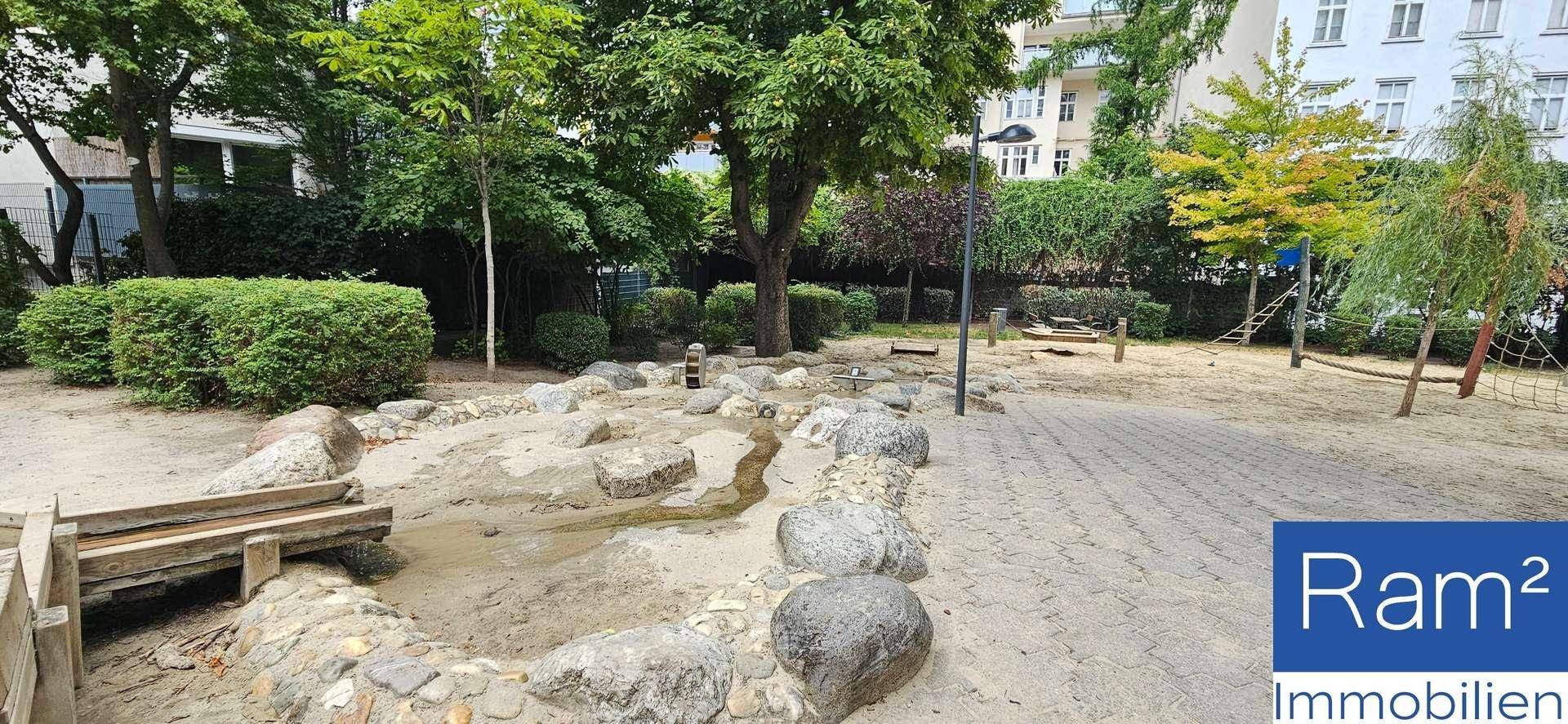 Spielplatz mit Sandfläche, Felsen und Holzelementen, umgeben von Bäumen und Büschen.