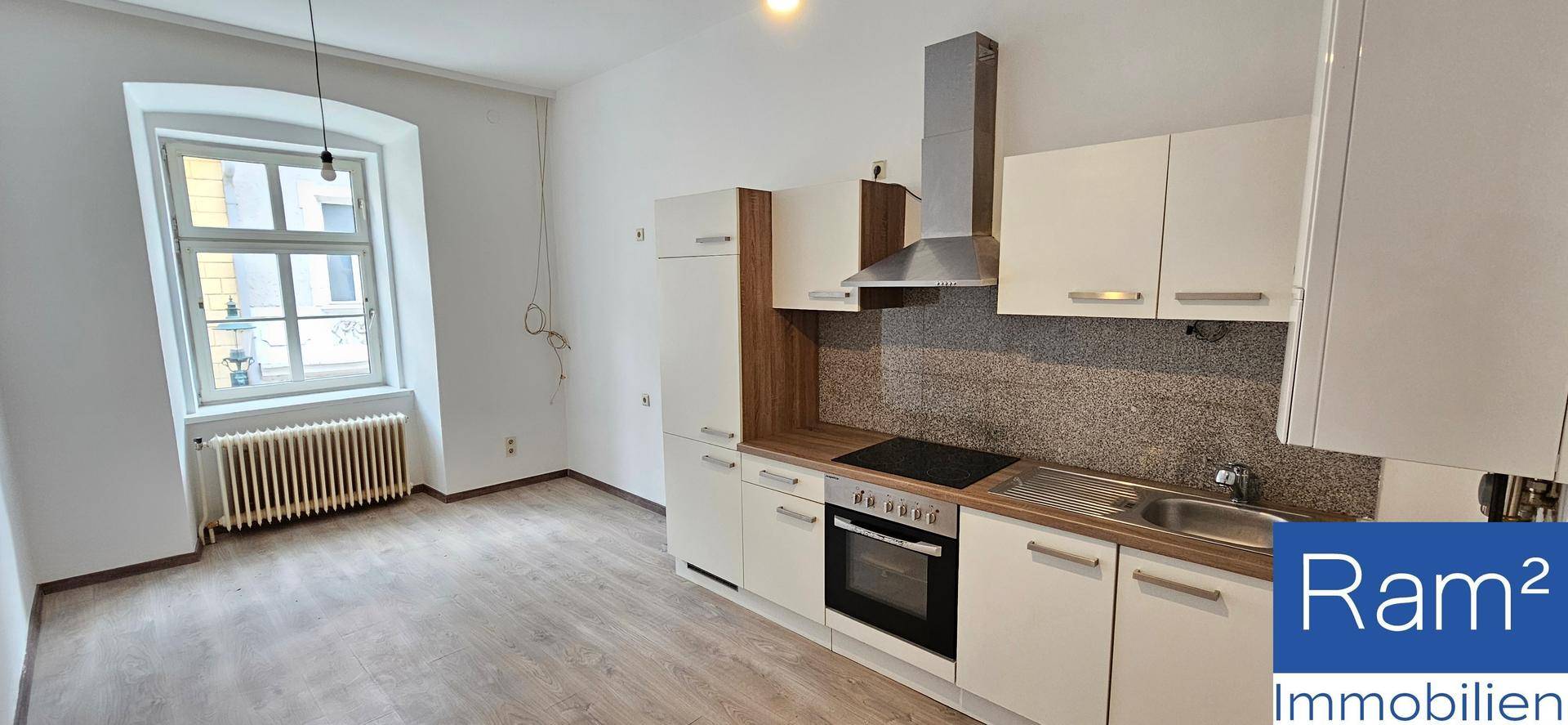 Helle Küche mit Einbauschränken, Kochfeld und Spüle, daneben ein Fenster mit Heizkörper.