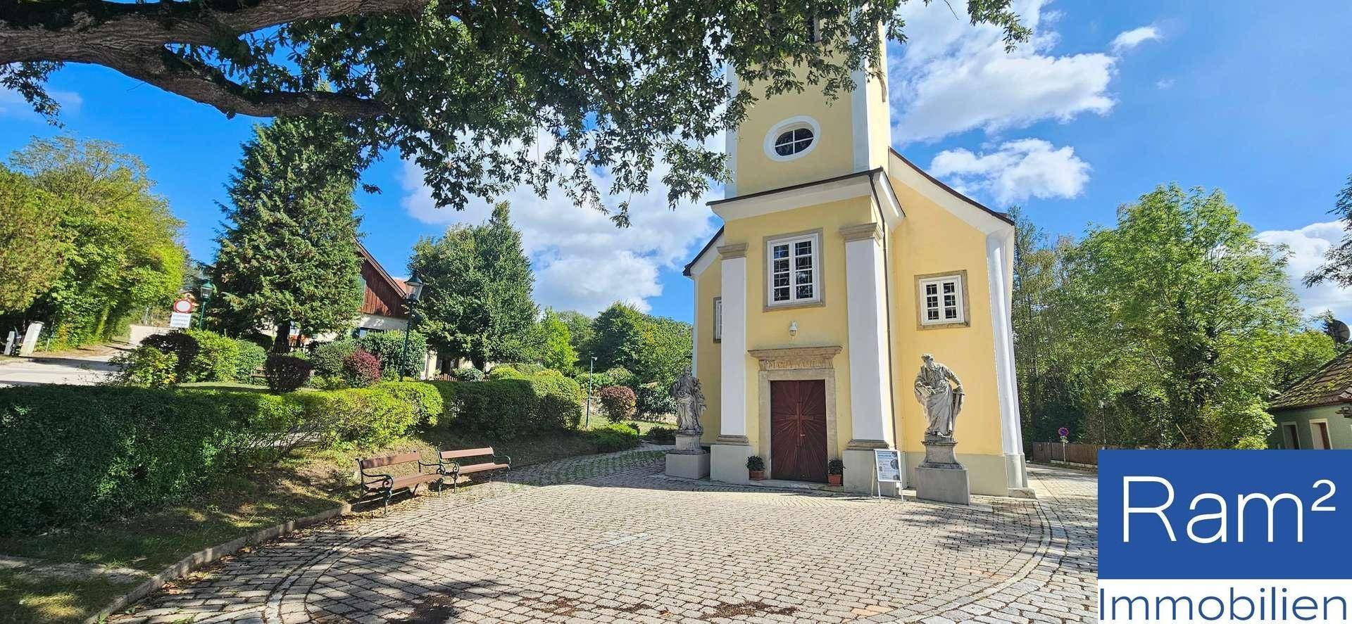 Kleine Kirche mit Bänken davor, umgeben von Bäumen und einem gepflasterten Vorplatz.