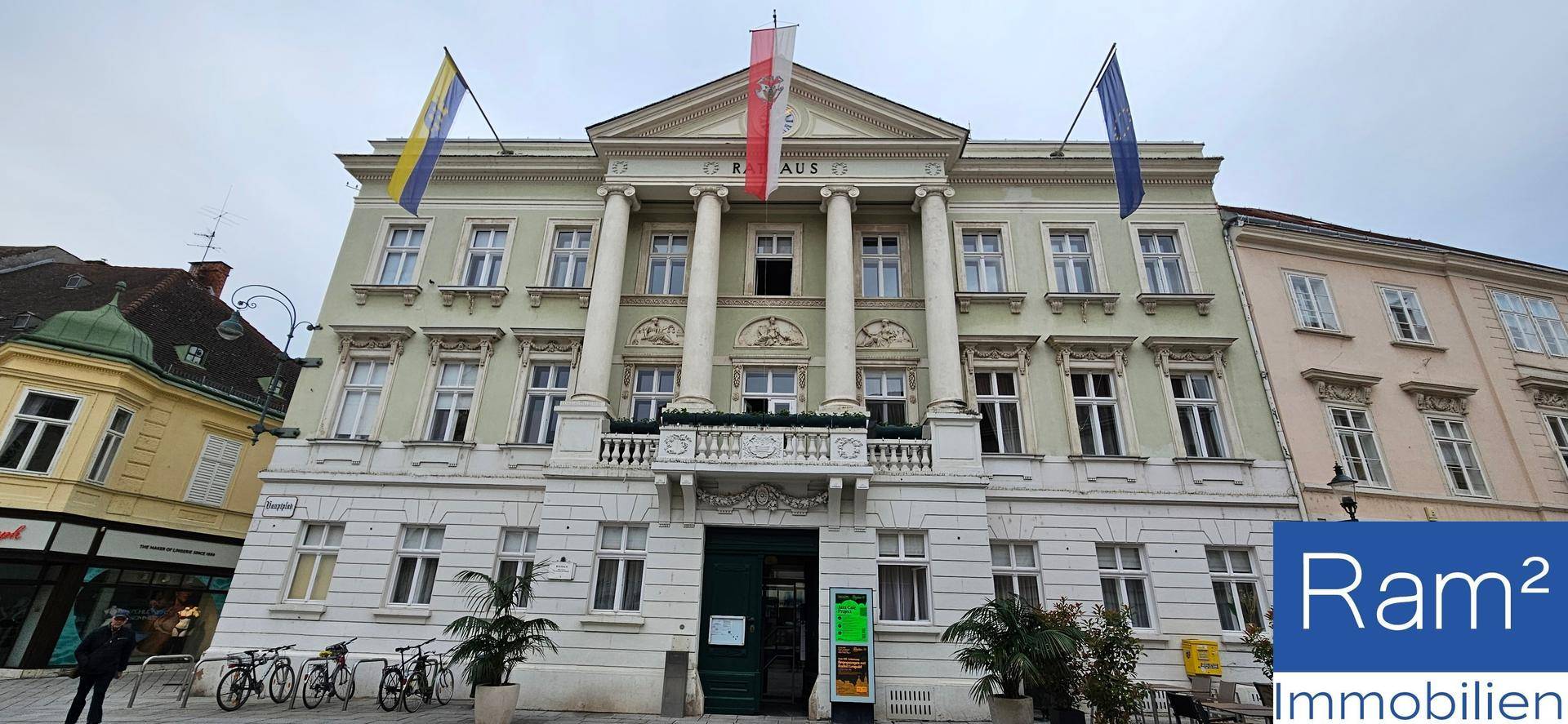 Repräsentatives Rathausgebäude mit Säulen und Balkon, flankiert von Fahnen unter bewölktem Himmel.