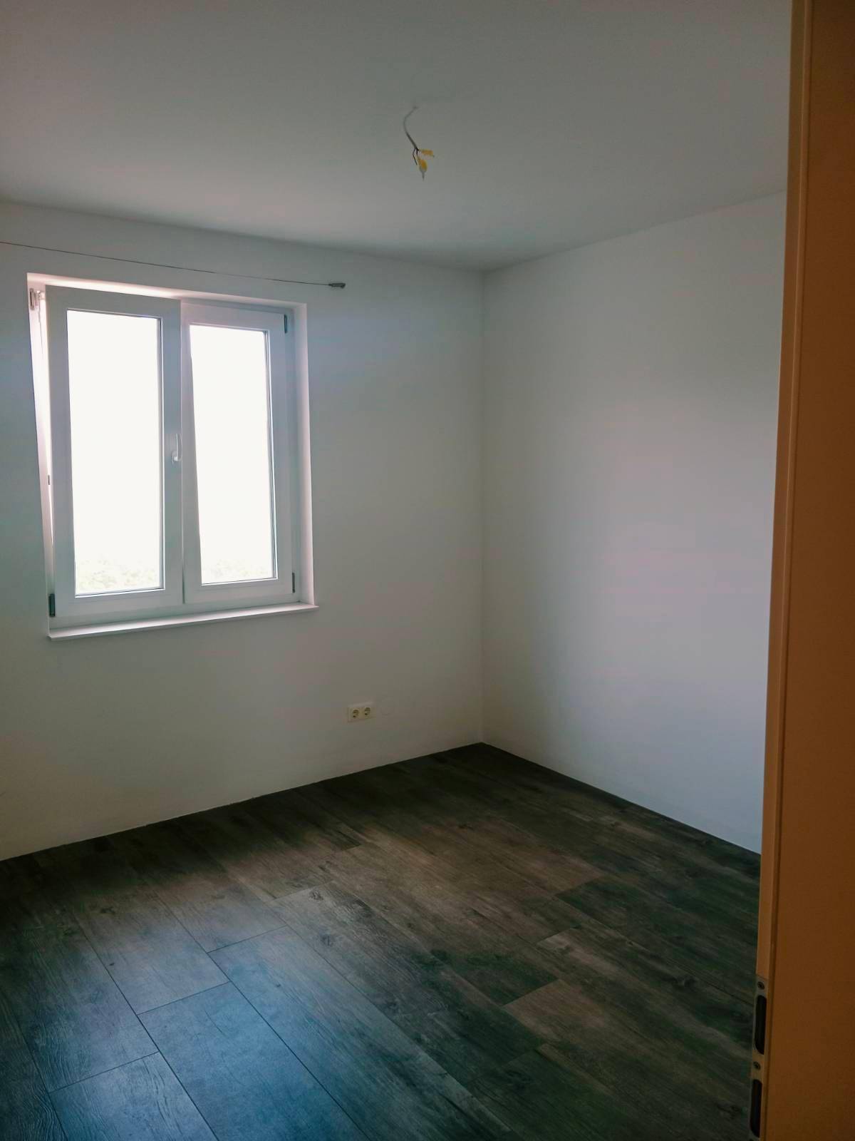 Leeres Zimmer mit dunklem Laminatboden und einem großen Fenster, bereit zur Einrichtung.