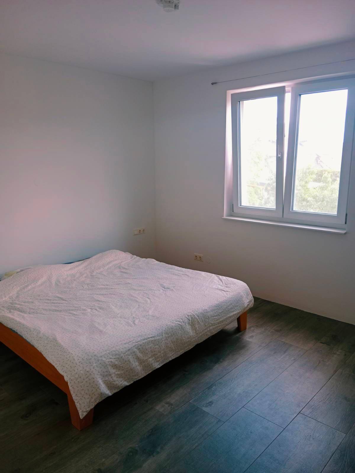 Einfaches Schlafzimmer mit einem Bett, dunklem Laminatboden und einem großen Fenster.