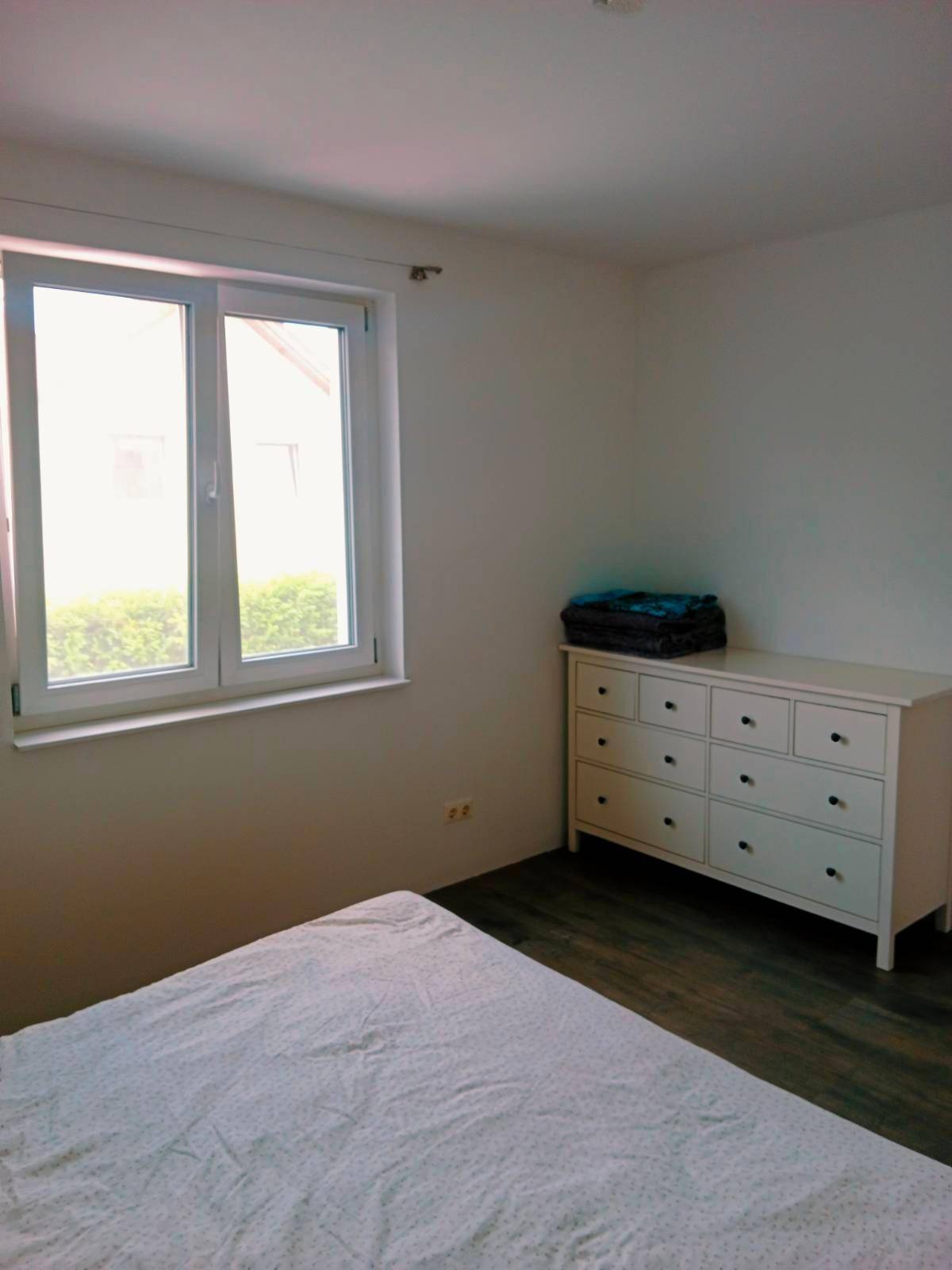 Helles Schlafzimmer mit einem Fenster, dunklem Laminatboden und einer weißen Kommode.