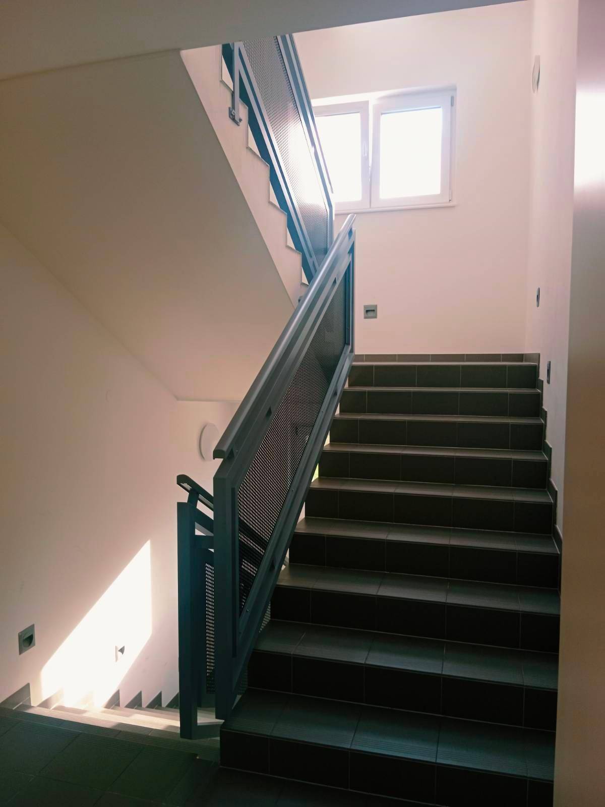 Helles Treppenhaus mit dunklen Fliesenstufen und einem Fenster für Tageslicht.