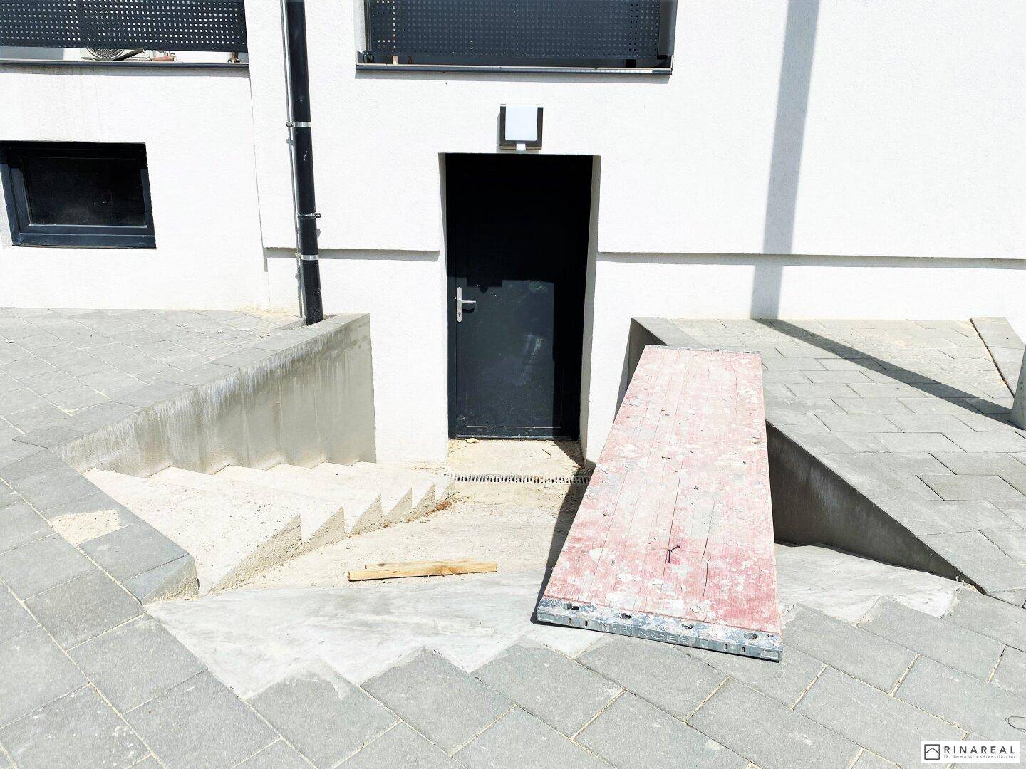 Eingangsbereich mit provisorischer Rampe, Betontreppe und dunkler Tür zu einem Untergeschoss.