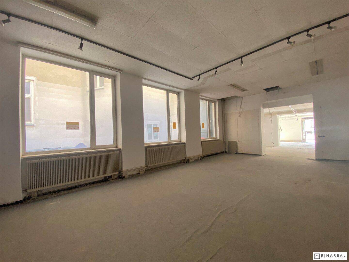Großer, leerer Raum mit mehreren Fenstern, Heizkörpern und Betonboden, ausgestattet mit Schienenbeleuchtung.