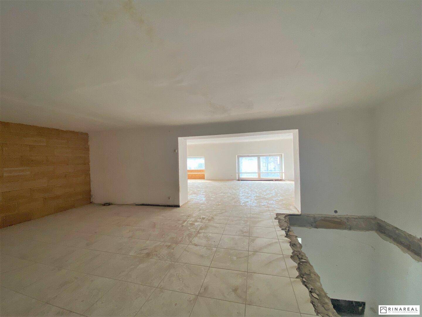 Heller Raum mit Fliesenboden und Holzwand, offen zu einem weiteren Bereich mit Fenstern und Tageslicht.