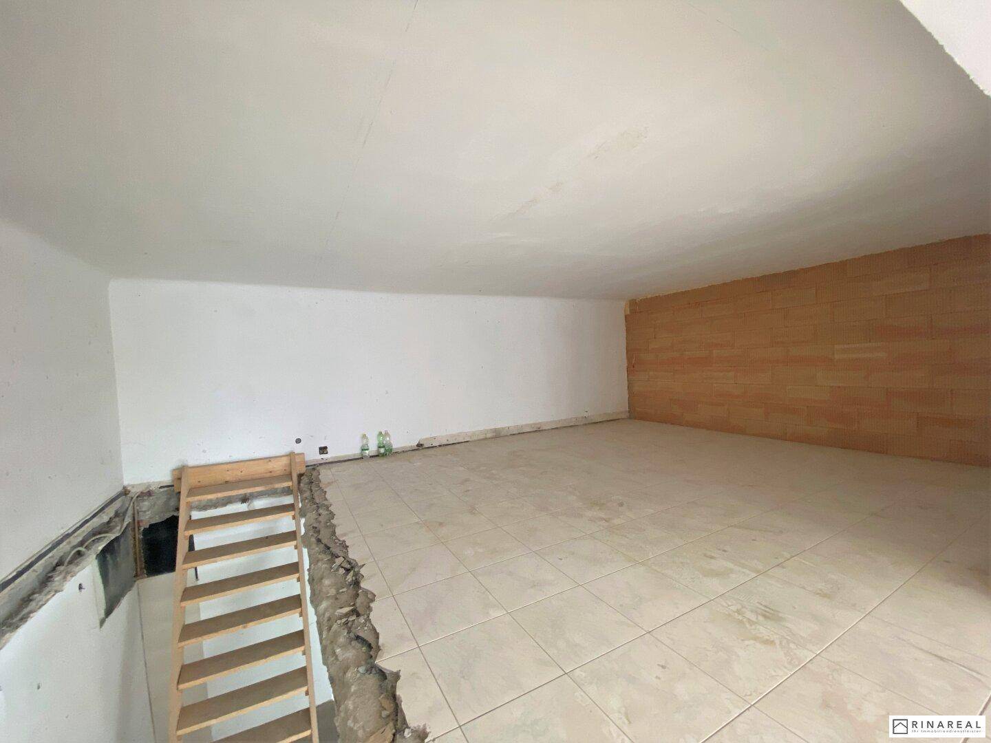 Innenraum mit hellen Fliesen, einer Holzakzentwand und einer Treppe zu einer unteren, noch unfertigen Ebene.