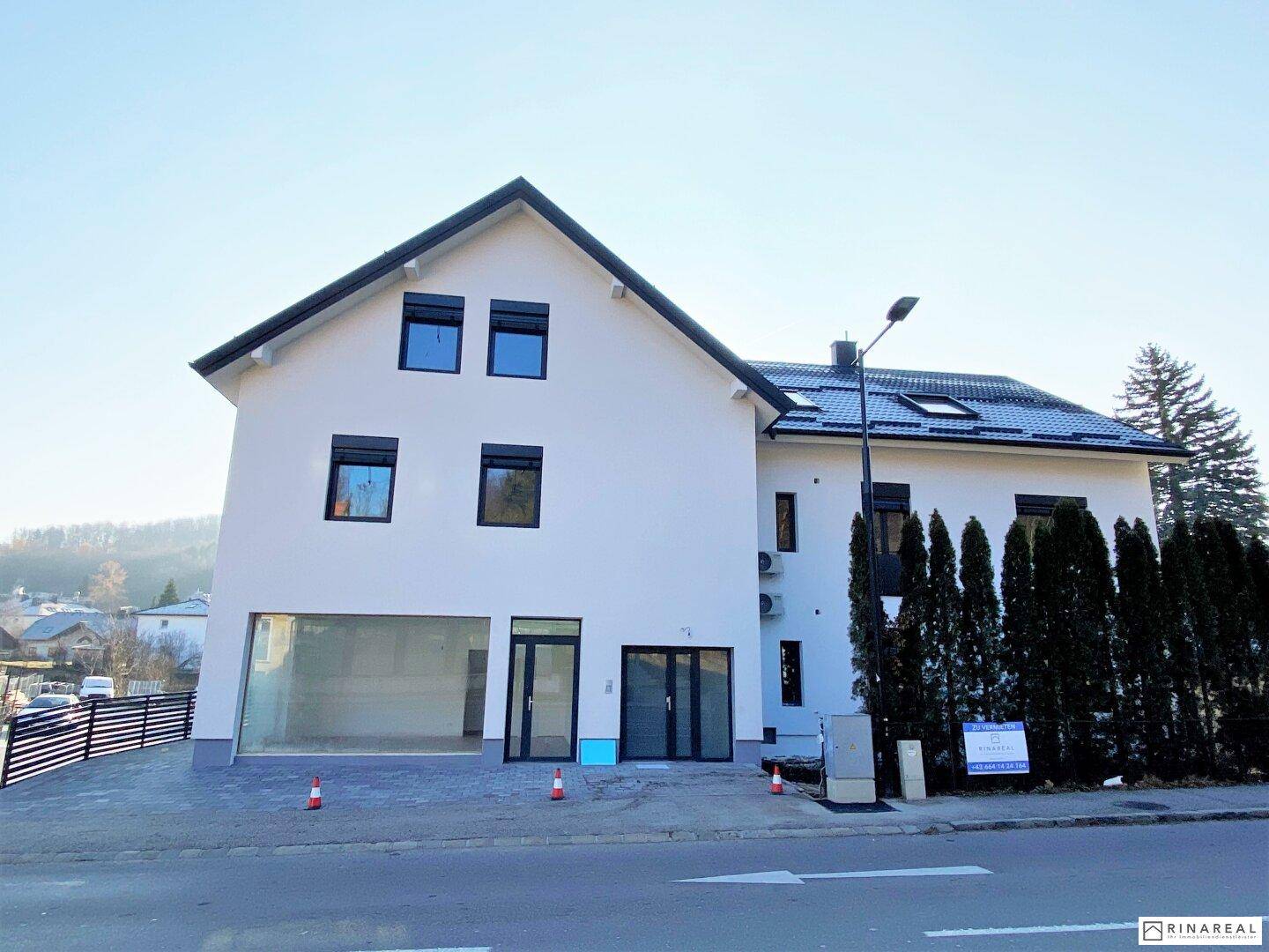Modernes Gebäude mit weißer Fassade, großen Fenstern und Rollläden an einer Straße gelegen.