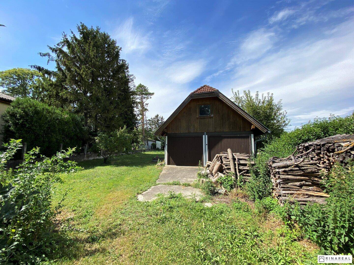 Rustikales Holzgebäude mit zwei Garagentoren und Holzstapeln in einem grünen Garten.