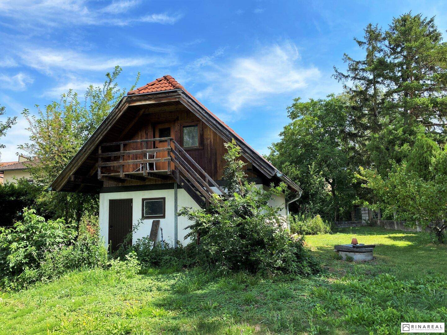 Kleines Holzhaus mit weißem Sockel, externer Treppe und Balkon in einer grünen Umgebung.