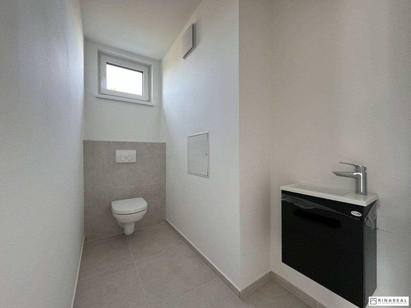 Separates Gäste-WC mit Wand-WC, kleinem Waschbecken und Fenster für natürliche Belüftung, sauber und praktisch.