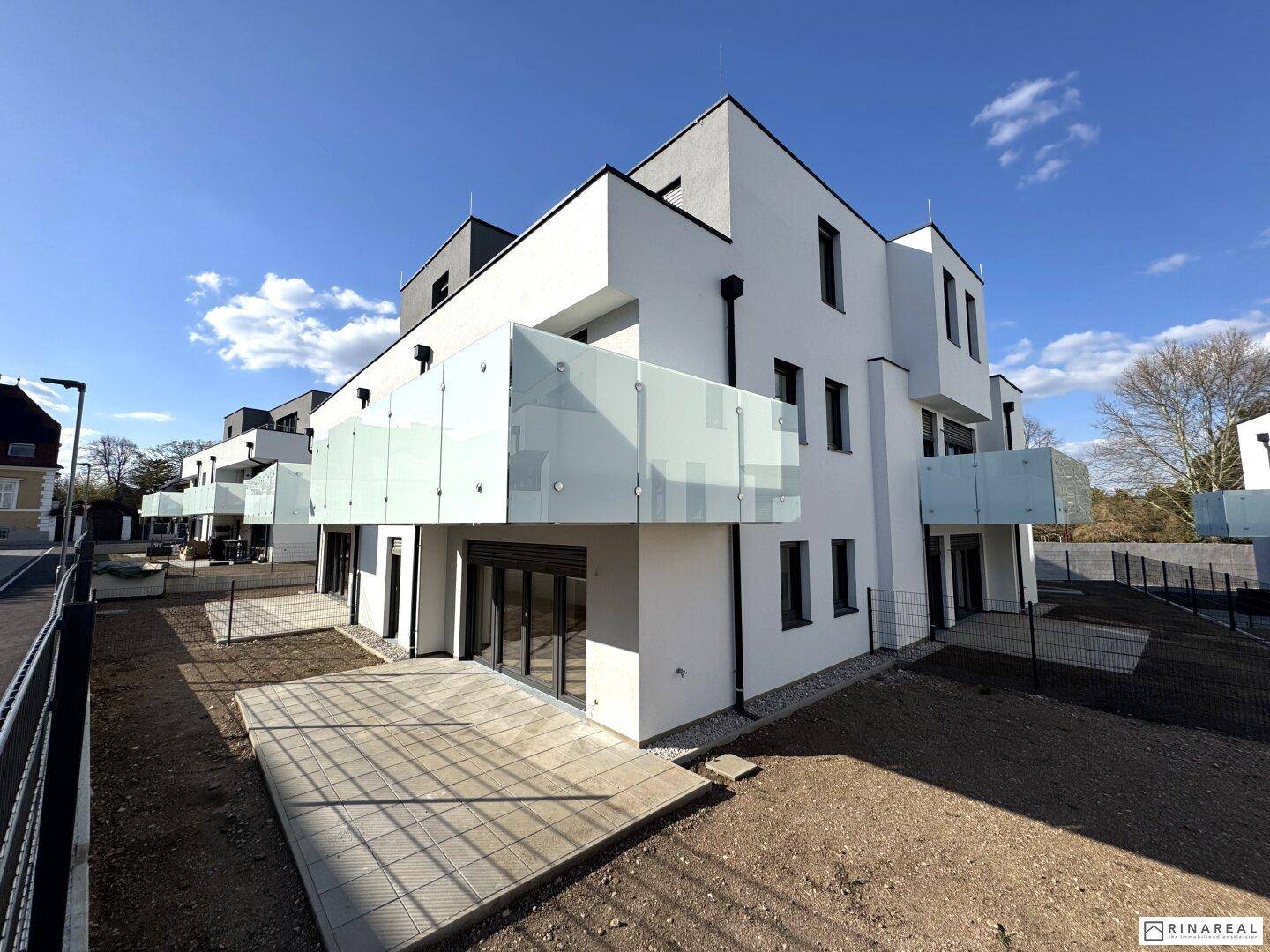 Modernes Mehrfamilienhaus mit weißer Fassade, Glasbalkonen und Terrassen im Erdgeschoss, Neubau.
