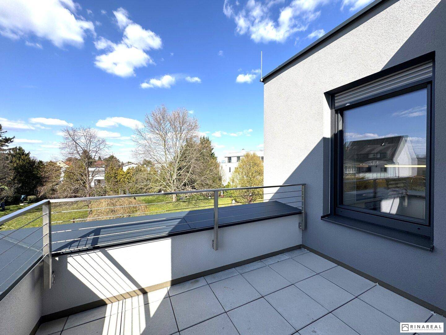 Sonniger Balkon mit Fliesenboden und Blick ins Grüne sowie auf umliegende Gebäude an einem klaren Tag.