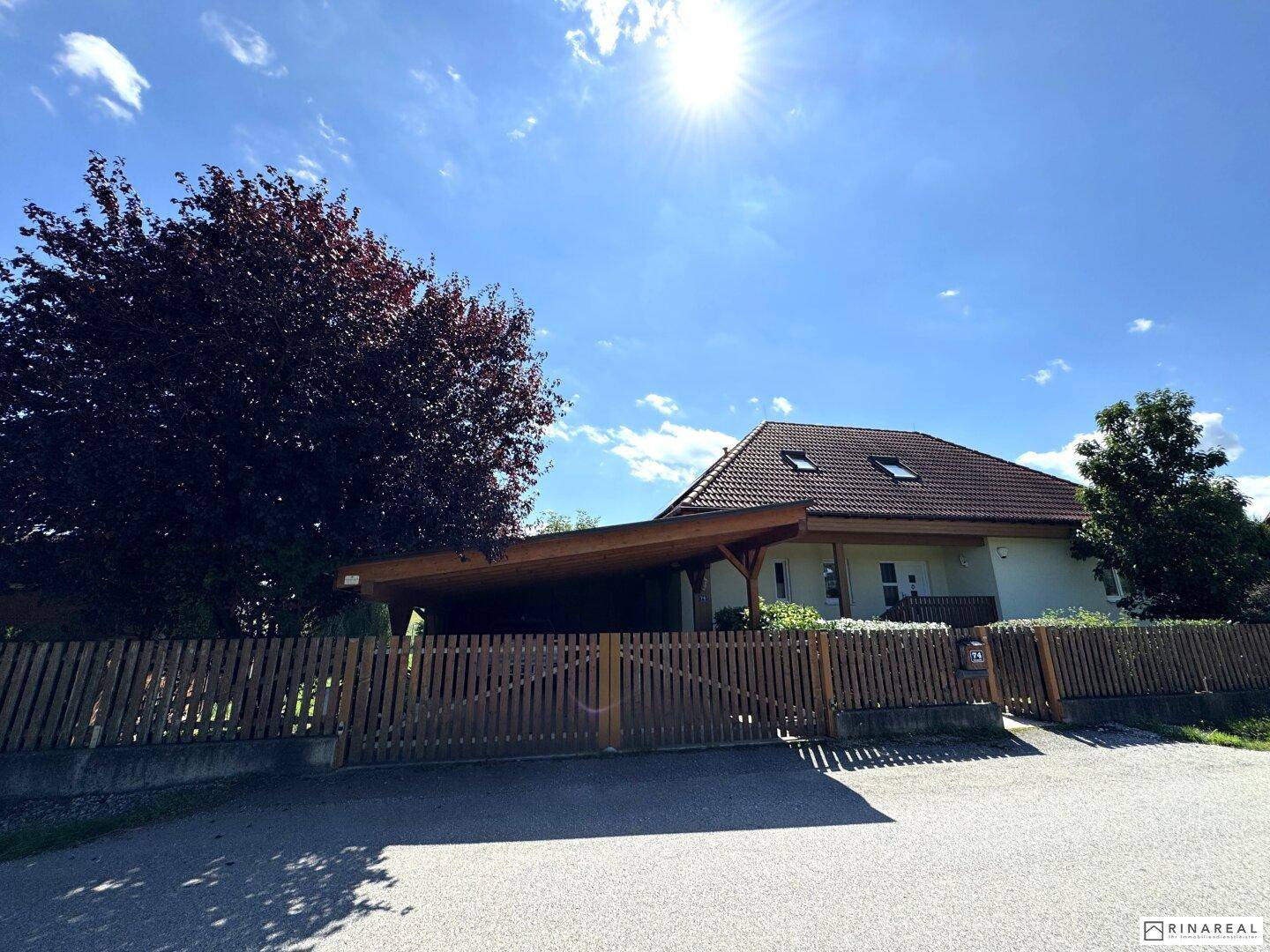 Hausfassade mit Carport und Holzzaun, umgeben von Bäumen unter blauem Himmel.