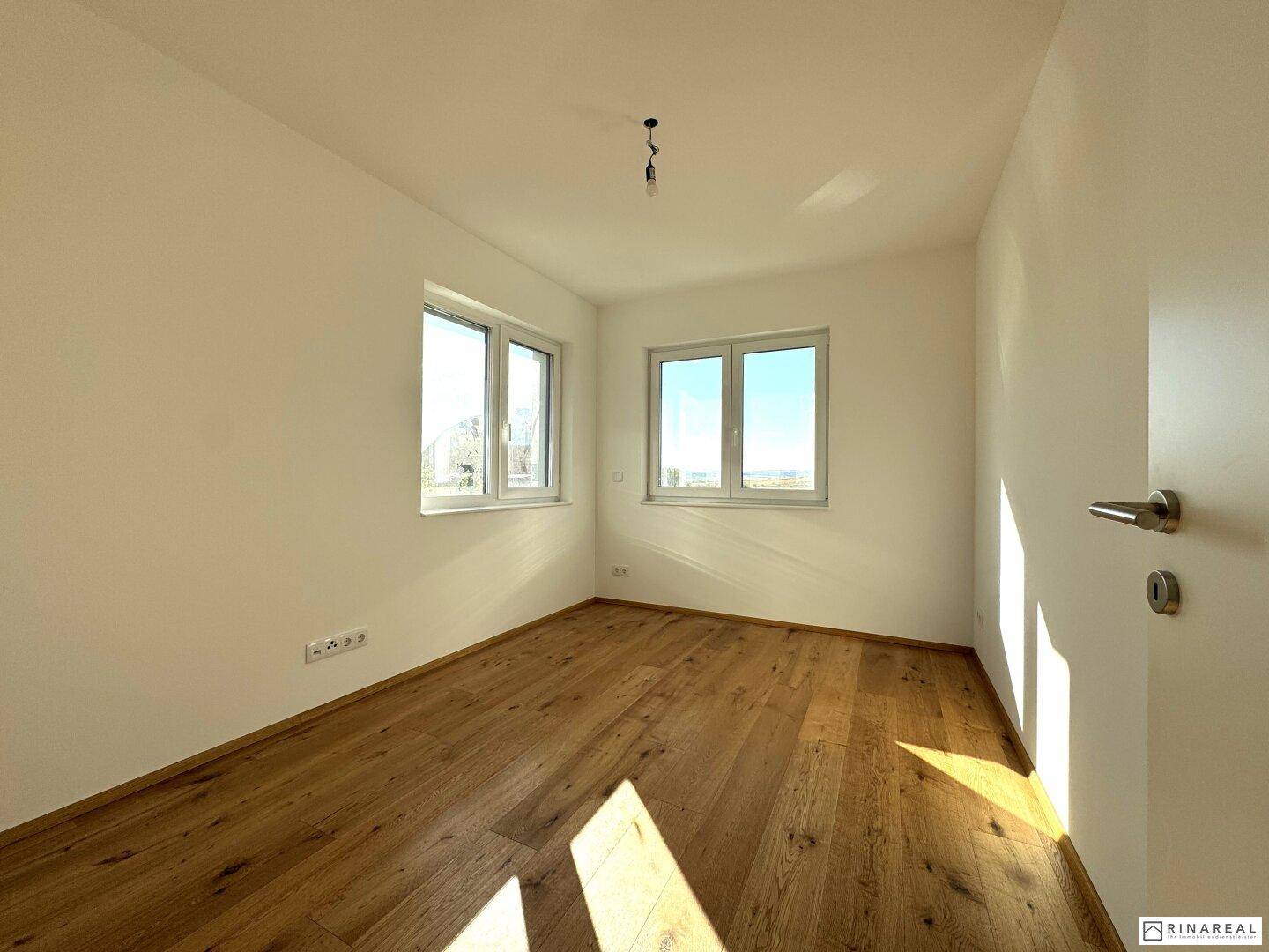 Helles Zimmer mit Holzboden und zwei Fenstern, die viel Sonnenlicht hereinlassen.